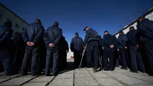 Чоловіки у темно-синій формі стоять у ряд з руками за спиною на подвірї з колючим дротом