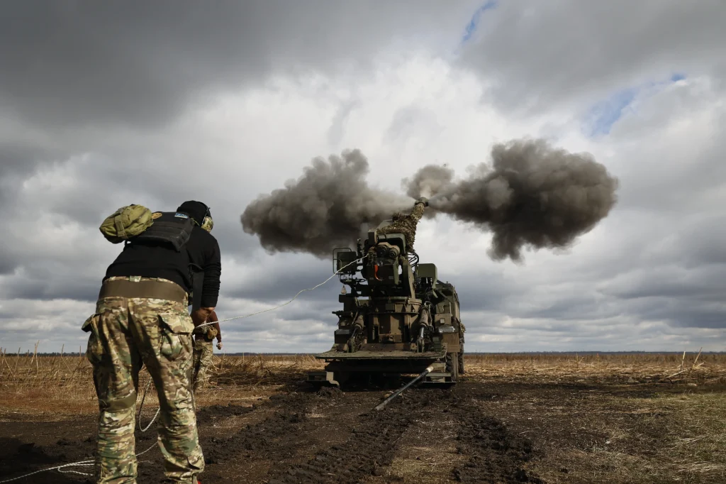 Військовослужбовець спостерігає постріл самохідної гаубиці на брудному полі під хмарами