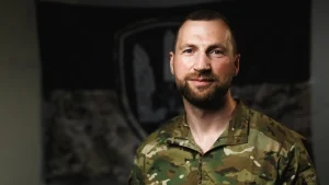 Чоловік з бородою у камуфляжній формі на тлі емблеми зі щитом