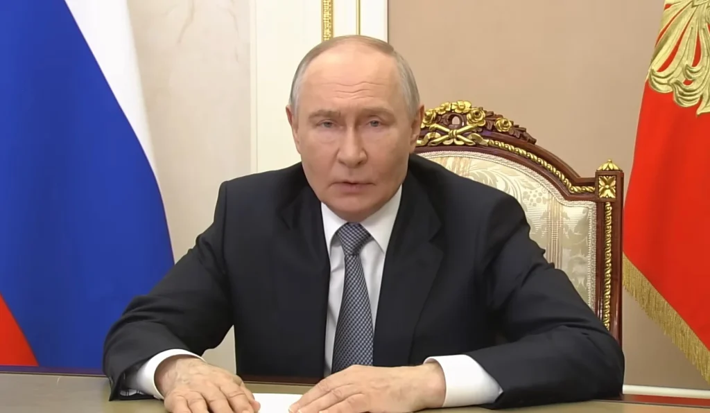Володимир Путін сидить за столом у чорному костюмі на тлі російських прапорів