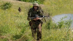 Військовий у камуфляжі з автоматом іде через високу траву біля водойми